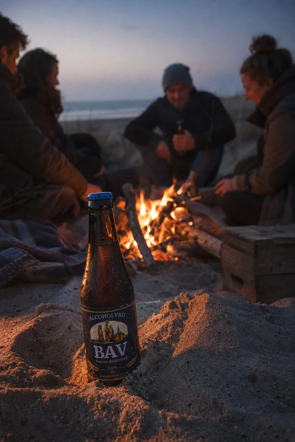 Vrienden rond een kampvuur in de duinen bij schemering, met een flesje BAV in het zand