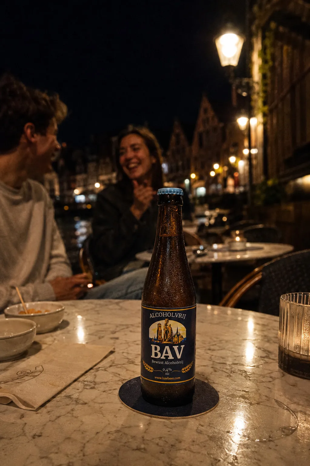 Twee vrienden lachen aan een cafétafel bij een Brugse gracht, met vooraan een flesje BAV op een bierviltje