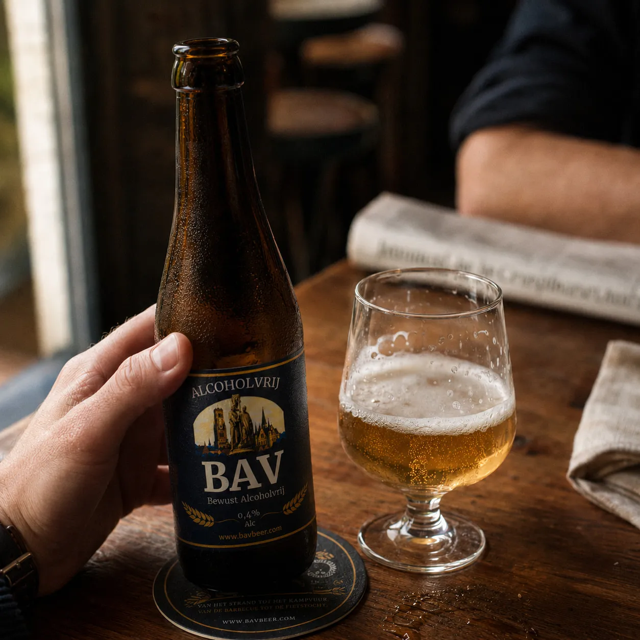 Een hand zet een flesje BAV op een bierviltje, naast een glas goudblond bier op een cafétafel